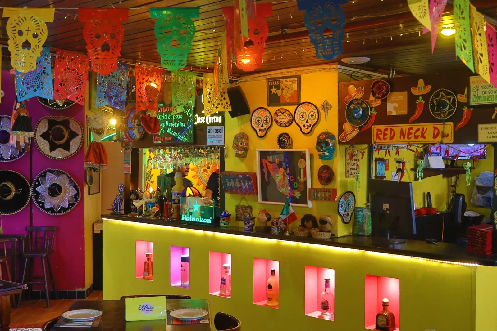 Detalhe do bar com bartender em ação, papel picado e garrafas iluminadas