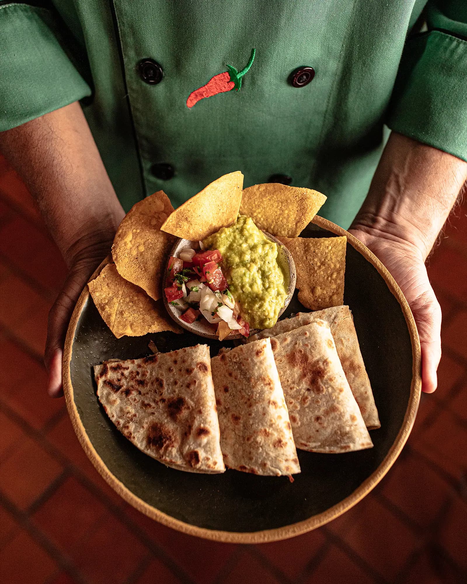 Chef de dólmã verde servindo prato de quesadillas com guacamole e pico de gallo