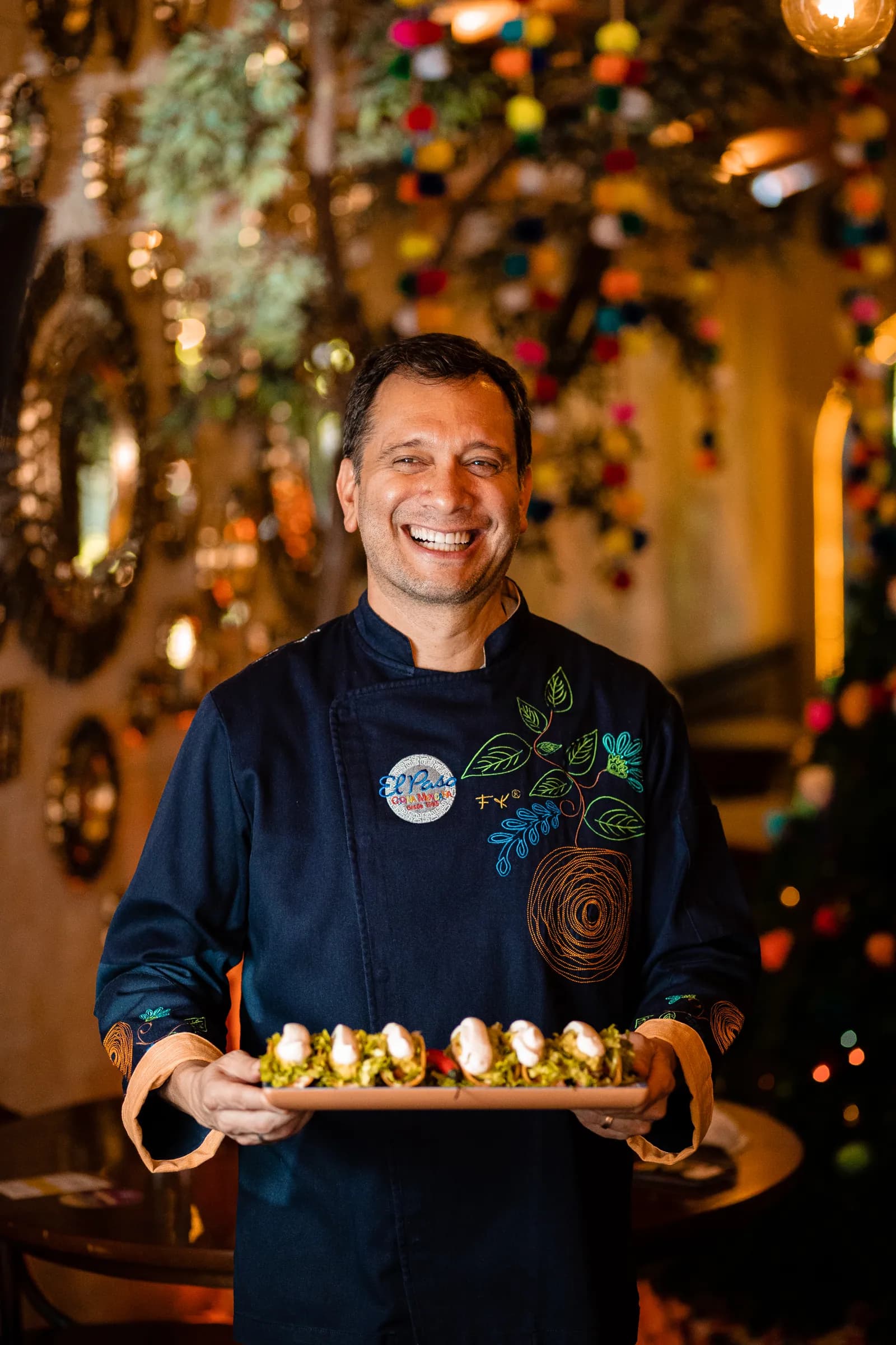 Chef sorridente segurando bandeja de tacos no salão decorado com espelhos e pompons