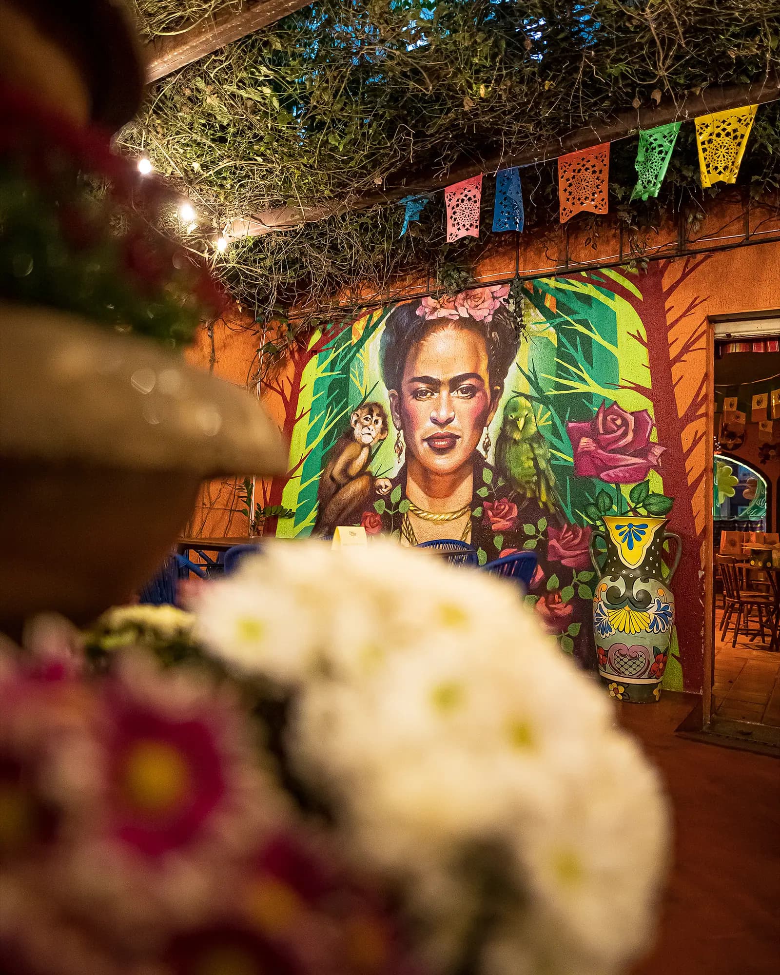 Mural da Frida Kahlo visto do pátio, com bandeirinhas de papel picado e flores em primeiro plano