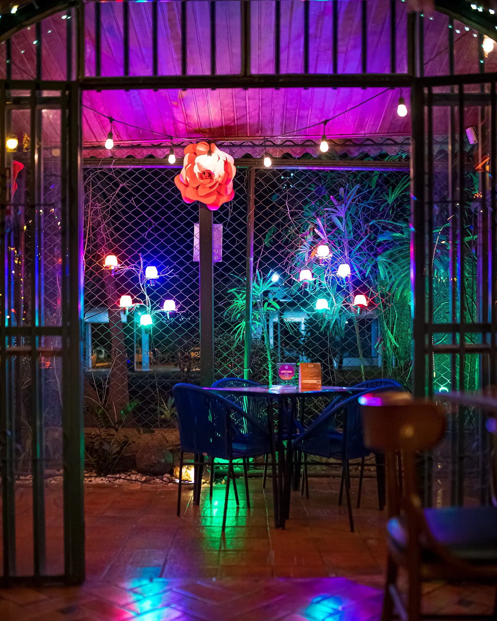 Jardim externo à noite com varal de lâmpadas, luzes coloridas e mesas azuis