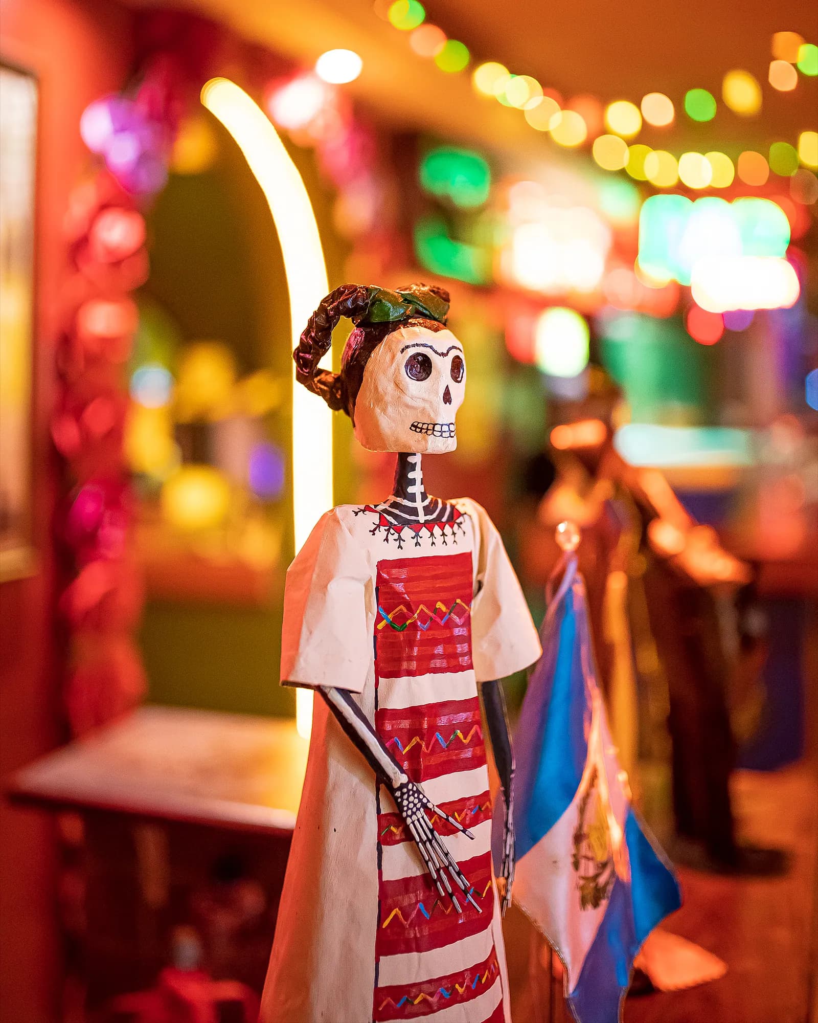 Catrina de papel machê com vestido bordado, entre luzes coloridas do bar