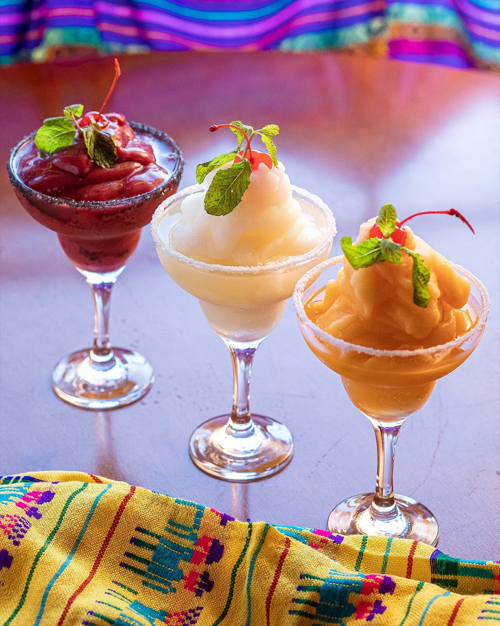 Três margaritas frozen de frutas sobre toalha mexicana colorida