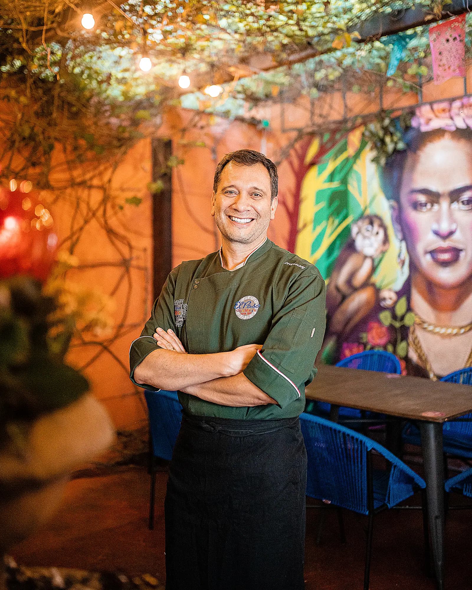 Chef sorridente de dólmã verde no pátio, diante do mural da Frida Kahlo