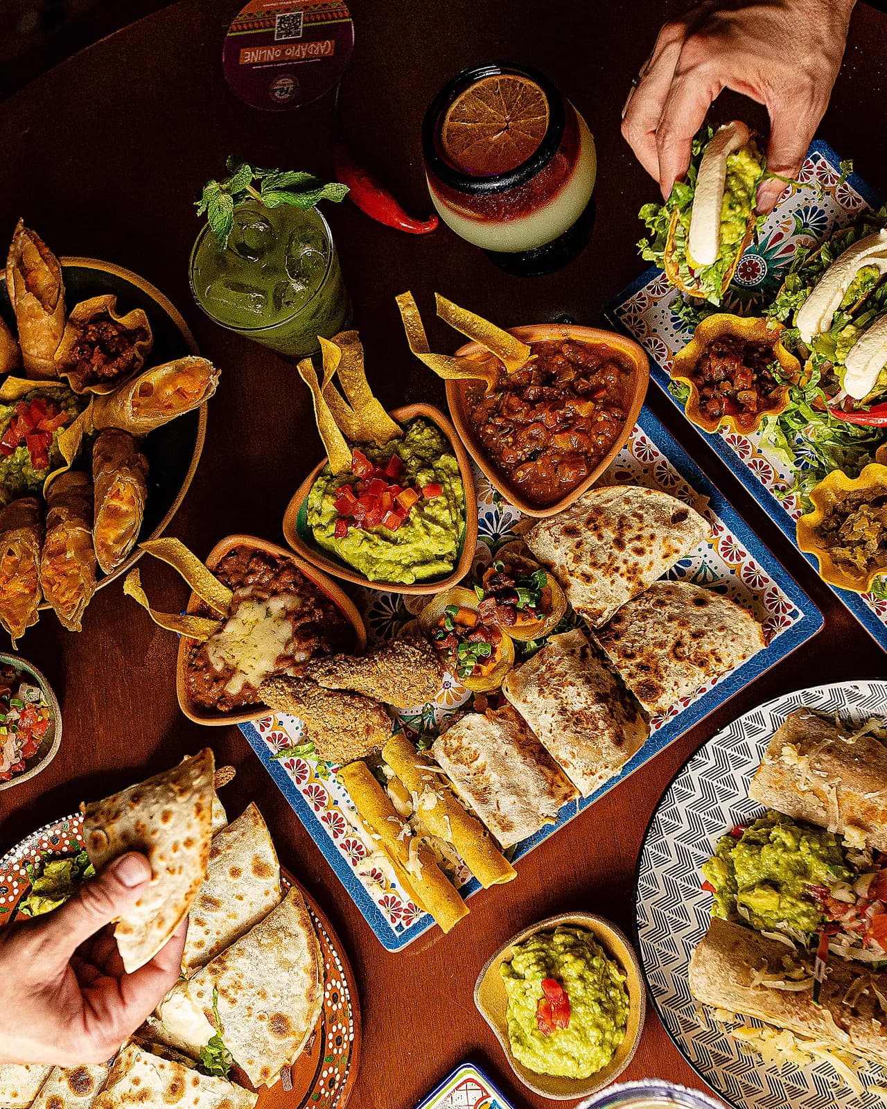 Mesa farta vista de cima: guacamole, quesadillas, flautas, margaritas e mãos se servindo